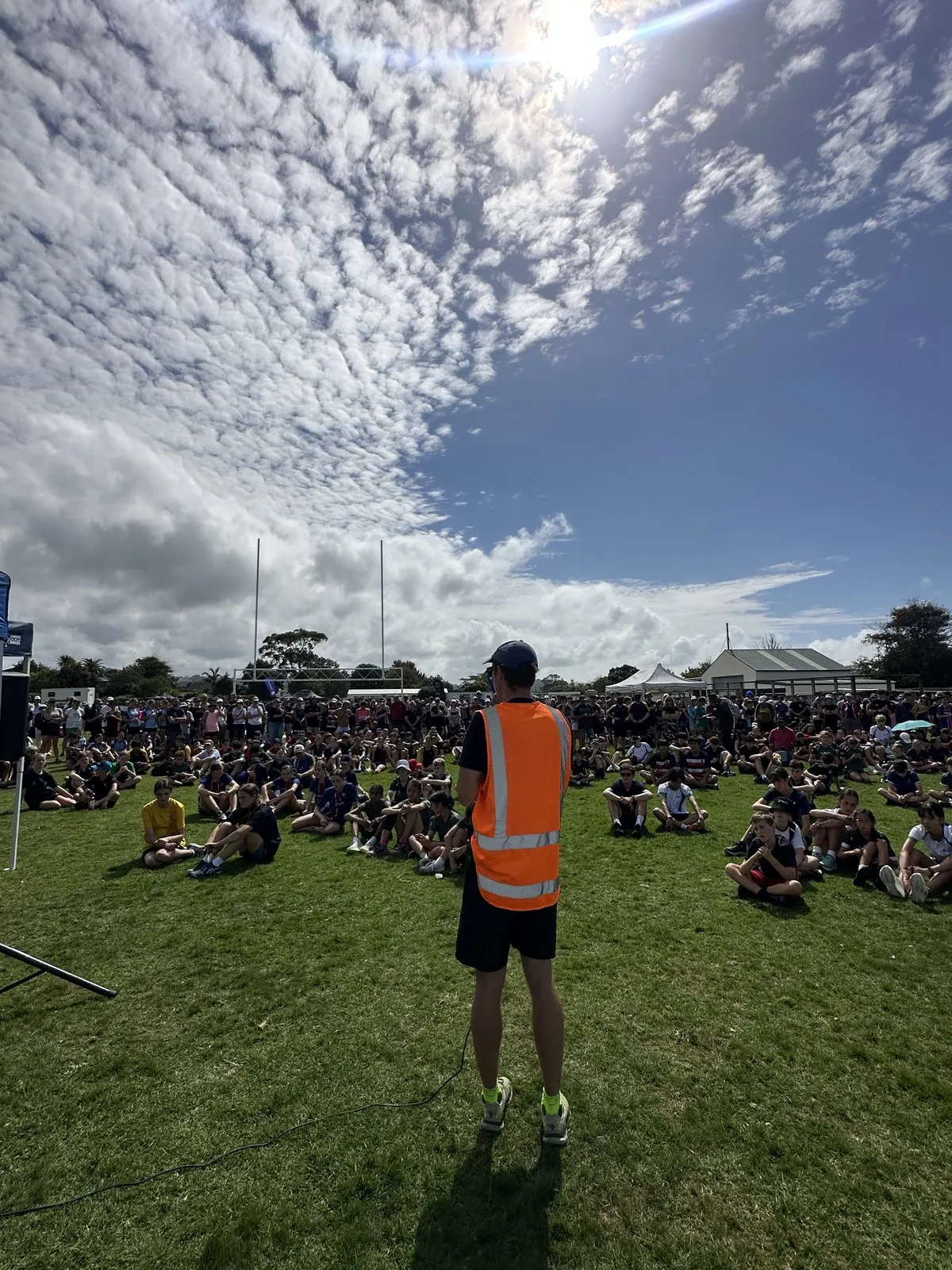 A TrailBlaze official in a hi-vis vest briefing several hundred students sitting on a school field at the Auckland Schools Sprint Series